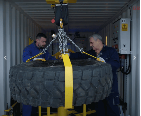 Technicians handling heavy military tire with overhead crane inside container workshop